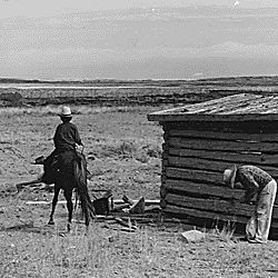 [Navajo man on a horse and another by a log cabin house on the Navajo reservation.]