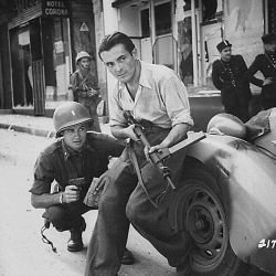 American officer and French partisan crouch behind an auto during a street fight in a French city.