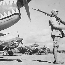 A Chinese soldier guards a line of American P-40 fighter planes, painted with the shark-face emblem of the "Flying Tigers," at a flying field somewhere in China. The American pursuit planes have a 12-